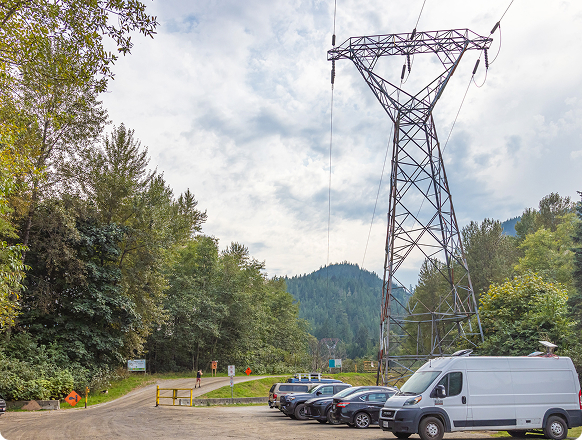 Fisherman's Park, Squamish, B.C. – A dirt road leading into a treed area with a large metal electricity tower prominently featured. A few parked cars and a white van are on the right, and distant mountains are visible under a cloudy sky.