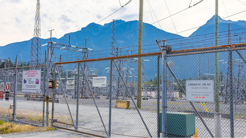 Cheekye Substation – A chain-link fence with barbed wire at the top, enclosing an electrical substation. Several signs are attached to the fence, mountains are visible in the background.