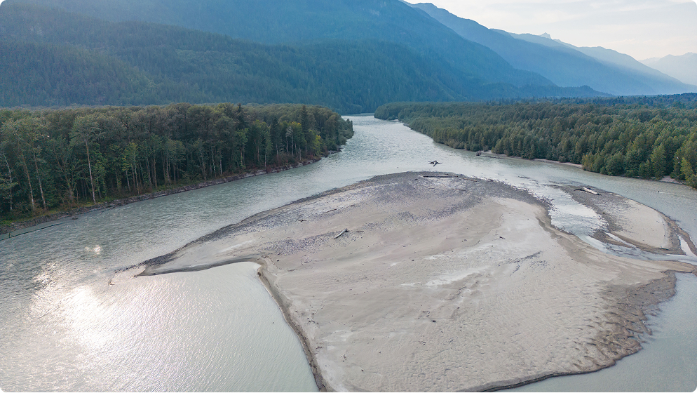 Squamish River, Squamish, B.C – An aerial view of a wide river splitting around a large, light-colored sand and gravel bar. Dense green forests line both banks of the river, and mountains are visible in the background under a soft sky.